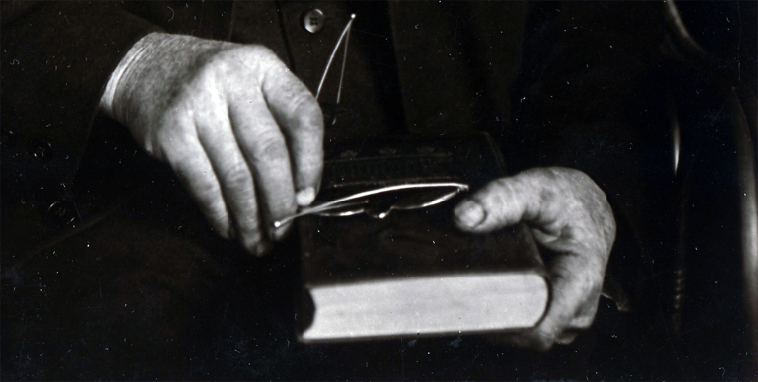 Two worn hands hold a book on a man's lap, with a pair of spectacles balanced atop the book